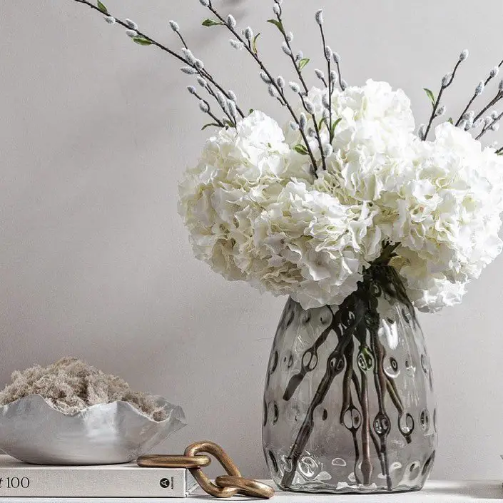 A vase filled with white hydrangeas sits beside an open book on a wooden table, creating a serene atmosphere.