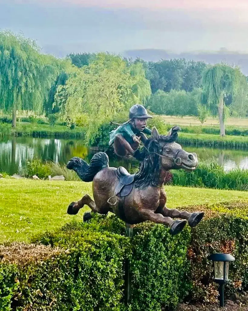 A metal statue depicting a horse and rider gracefully leaping over a hedge, showcasing dynamic movement and artistry.